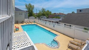 A rectangular swimming pool with a white deck and lounge chairs, surrounded by a white fence and houses in the background.