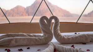 A room with a large window, a bed with white towels arranged in a heart shape, and scattered rose petals on the floor. - Wadi Rum Desert Camp (Wadi Rum)
