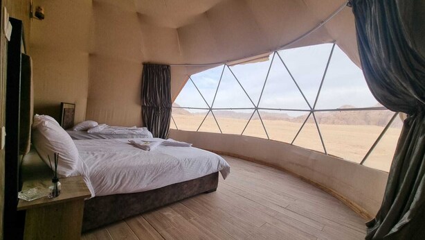 A bedroom with a large window offering a desert view, a bed with white linens, and a wooden floor. - Wadi Rum Desert Camp (Wadi Rum)