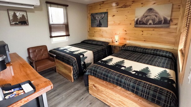 A cozy room with two beds, a desk, and a chair. There is a window with blinds, a wall-mounted air conditioner, and a lamp on a wooden table. - Escape to the tranquility of our cozy cabin inn, where rustic charm meets modern comfort. Experience a home away from home ambiance. (Spencerport)
