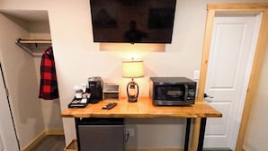 A compact kitchenette with a microwave, coffee maker, and a small refrigerator. A television is mounted above the counter, and a lamp is placed on the table. - Escape to the tranquility of our cozy cabin inn, where rustic charm meets modern comfort. Experience a home away from home ambiance. (Spencerport)