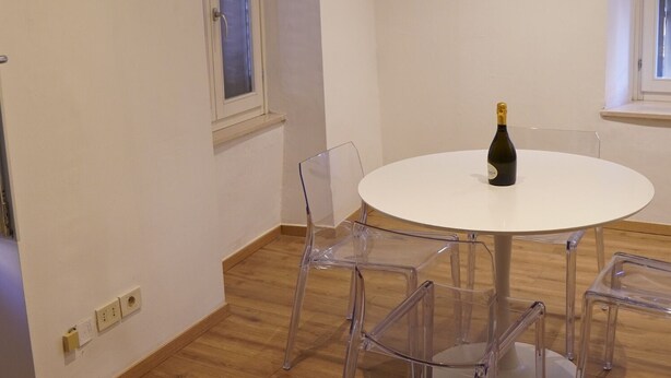 Een kamer met een ronde witte tafel, vier transparante plastic stoelen en een fles champagne op de tafel. Er zijn twee ramen met witte kaders en een deur met een witte omlijsting.