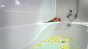 Une baignoire dans laquelle flottent des pétales de fleurs jaunes, une pomme de douche et une fleur rouge sur le mur.