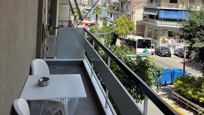 Un balcón con mesa y sillas, con vista a una calle concurrida, edificios y cielo despejado.