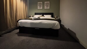 Una cama bien tendida con cabecera, una mesita de noche con lámpara y dos cuadros colgados en la pared.