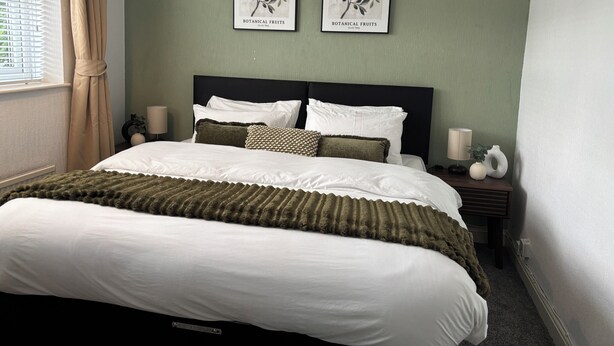 A neatly made bed with white and green bedding, two framed botanical prints on the wall, and a bedside table with a lamp and decorative items.