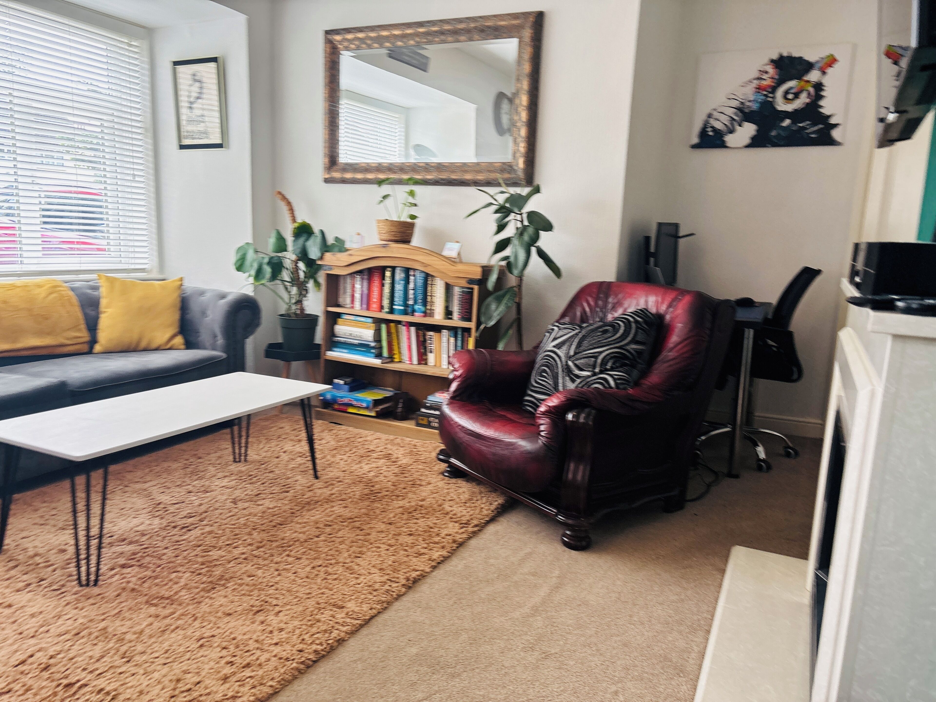 A living room with a sofa, a coffee table, a bookshelf, a red armchair, a mirror, and a potted plant.
