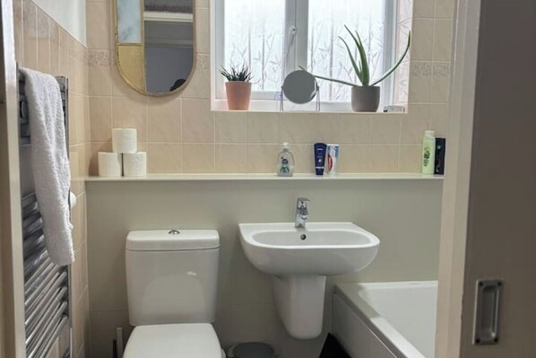 Un baño con inodoro, lavamanos y bañera. Hay toallas colgadas en un perchero, un espejo y plantas en macetas en el alféizar.