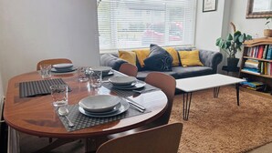 Un comedor con una mesa de madera dispuesta para cuatro, un sofá con cojines, una estantería y una ventana con persianas.