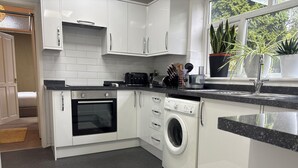 A modern kitchen with white cabinetry, a built-in oven, a washing machine, and a window with potted plants.