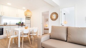 Una cocina moderna con armarios blancos, un comedor con mesa y sillas blancas, y un sofá beige en un espacio luminoso.