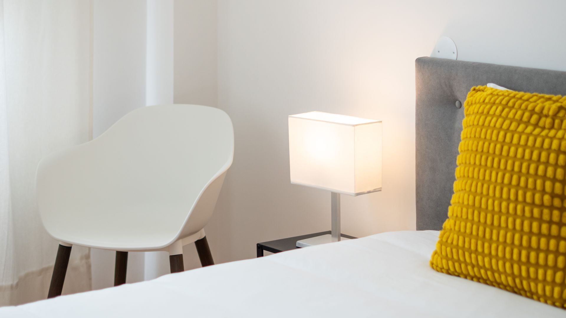 Une chambre d’hôtel moderne avec un lit blanc, une chaise blanche, une table de chevet avec une lampe et un oreiller jaune.