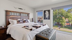 Un dormitorio con una cama grande, cabecera de madera, ventana con vista a un caballo y un pequeño banco.