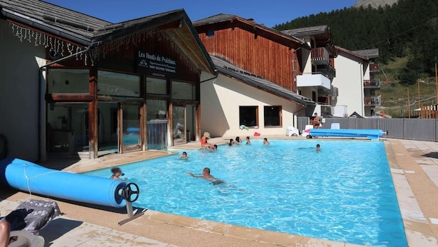 A swimming pool area with people swimming and lounging, a wooden building with a sign, and a mountain in the background. - Ski-in/ski-out & foot of the slopes, 2nd floor, swimming pool, sauna, balcony, tv, ski locker, 35m² (Les Orres)