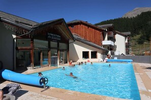 A swimming pool area with people swimming and lounging, a wooden building with a sign, and a mountain in the background.