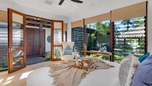 A modern bedroom with a large bed, a chair, and a view of tropical vegetation through large windows.