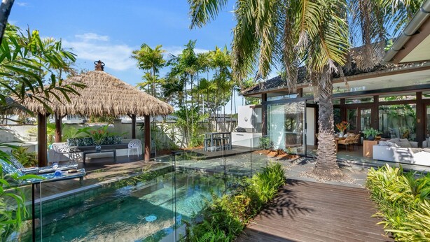 A modern outdoor pool area with a glass enclosure, wooden deck, and tropical plants.