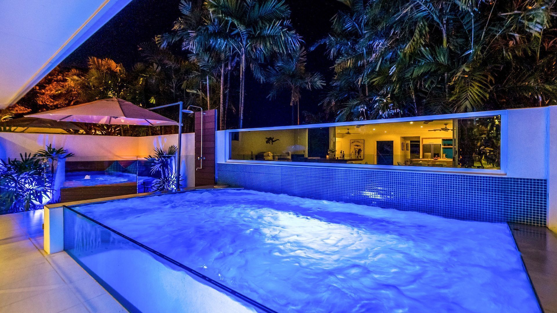 A modern outdoor pool area with a glass enclosure, palm trees, and a well-lit patio.