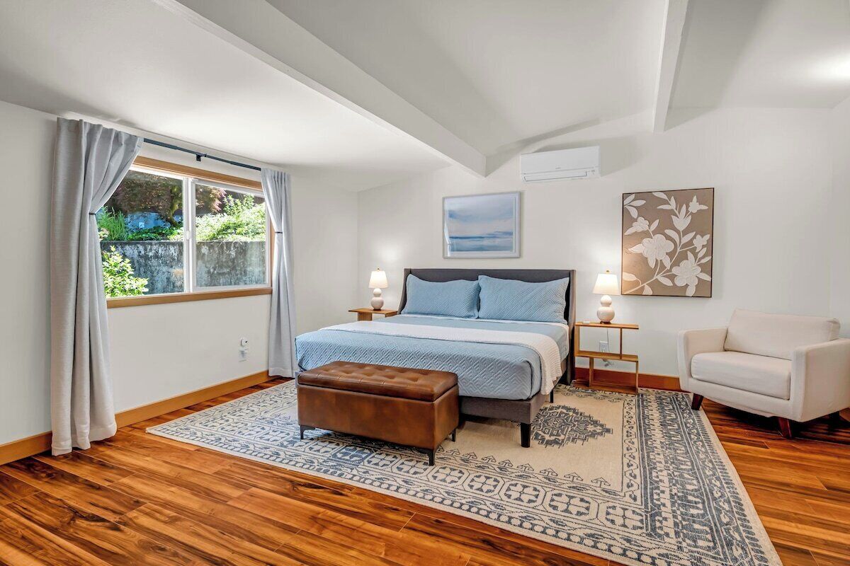 A bedroom with a large bed, a window with curtains, a patterned rug, a chair, and a wall-mounted artwork.