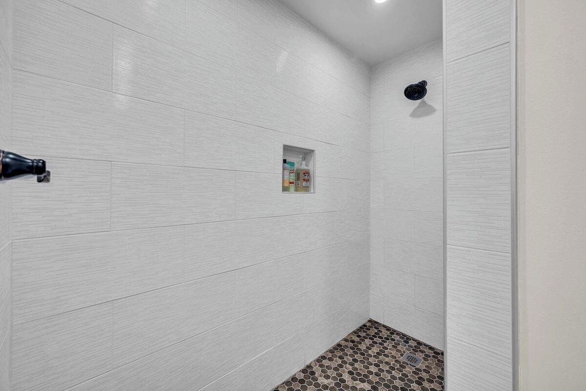 A bathroom with white tiled walls, a shower area, and a small shelf with toiletries.