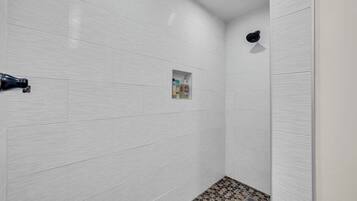 A bathroom with white tiled walls, a shower area, and a small shelf with toiletries.
