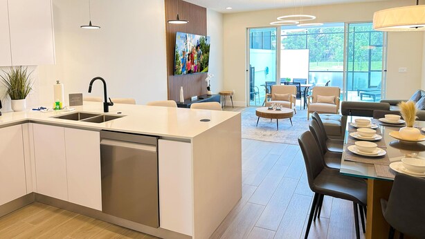 A modern kitchen with a dishwasher, a dining area with a glass table, and a TV mounted on the wall. - TR17406SB - New 3BR Villa w/ Pool | 17min Disney (Clermont)