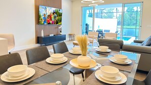 A modern dining area with a large TV, a dining table set for six, and a view of the outdoors through large windows. - TR17406SB - New 3BR Villa w/ Pool | 17min Disney (Clermont)