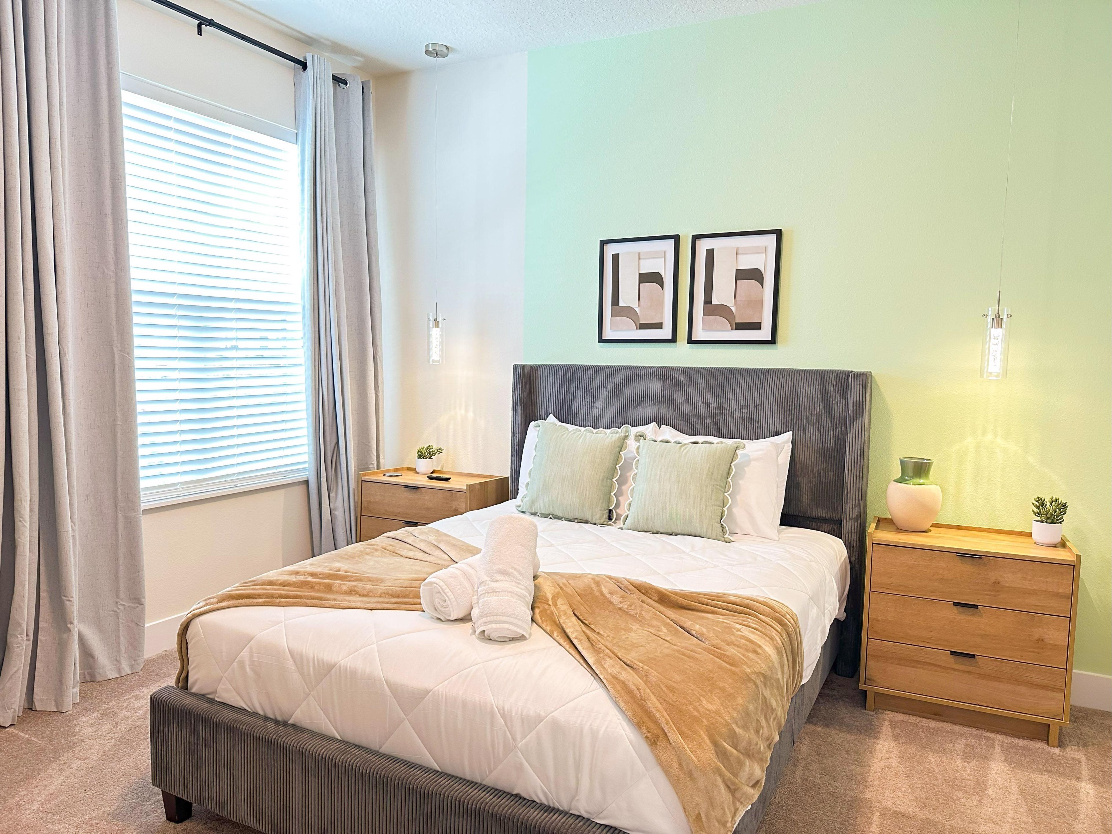 A neatly made bed with a brown blanket, a bedside table with a vase and a plant, and a wall with two framed pictures.