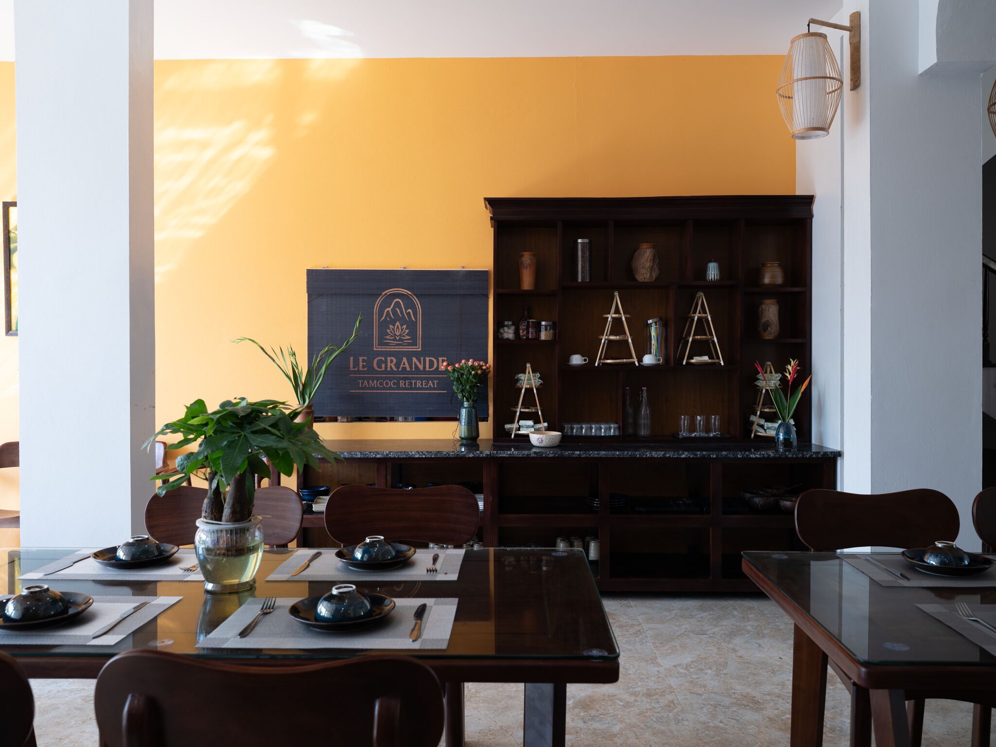A dining area with a table set for two, a potted plant, and a sign reading "Le Grande TAMCO Retreat".