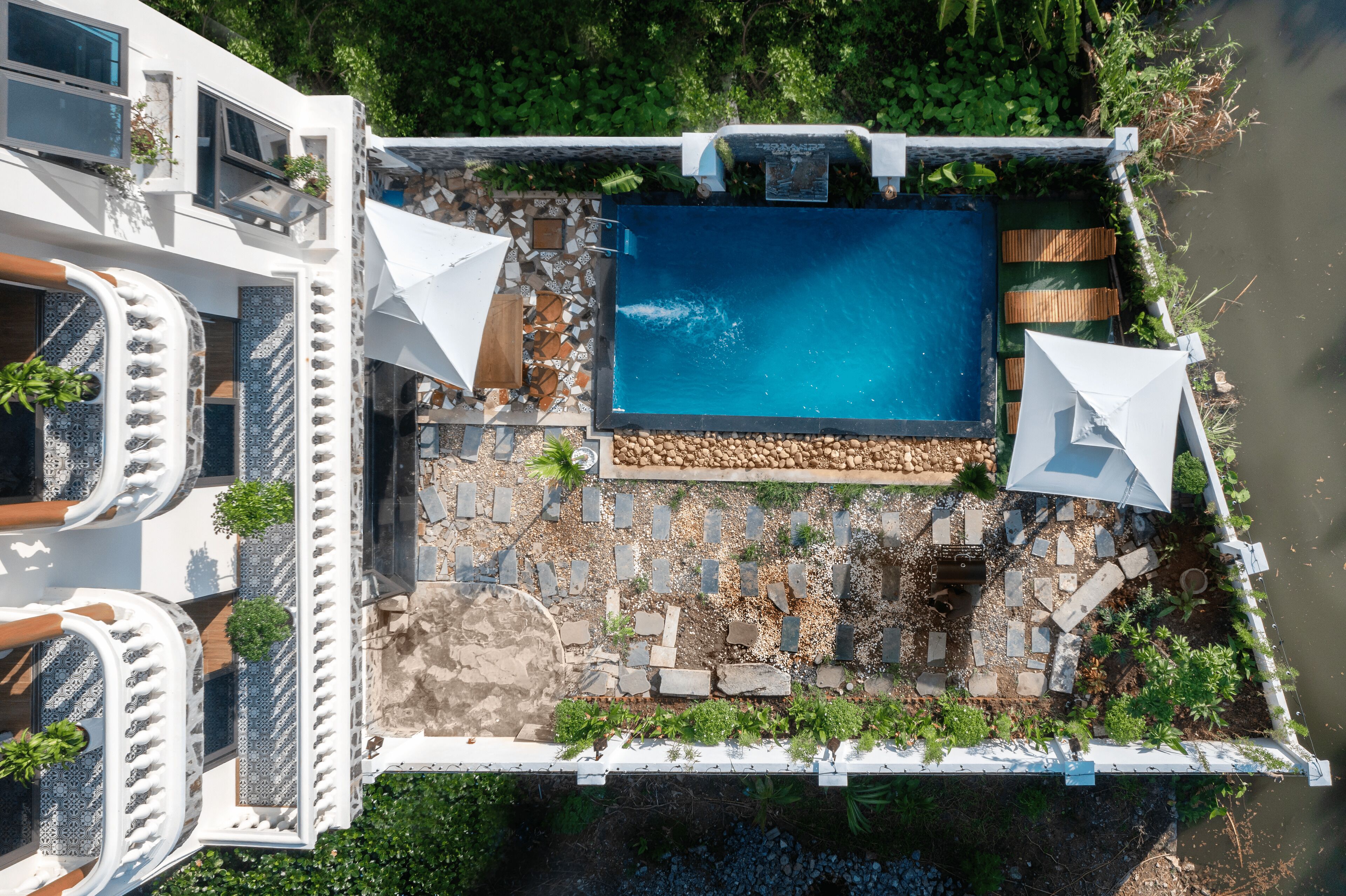 An aerial view of a modern pool area with a rectangular pool, surrounded by stone tiles and plants, and adjacent to a white building with balconies and windows.