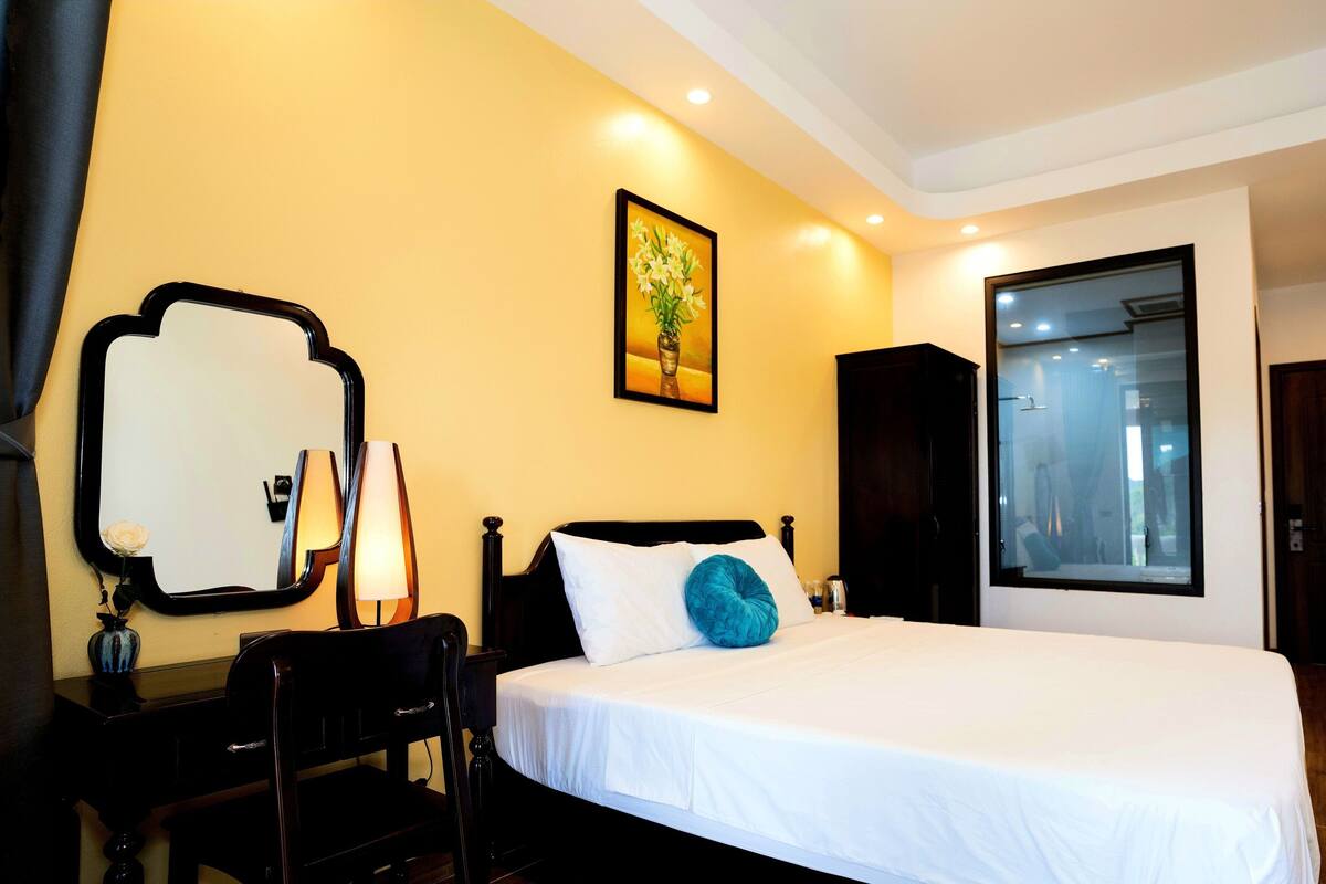 Une chambre d’hôtel avec un grand lit, un miroir, un tableau, une armoire et une salle de bain visible à travers une porte en verre.