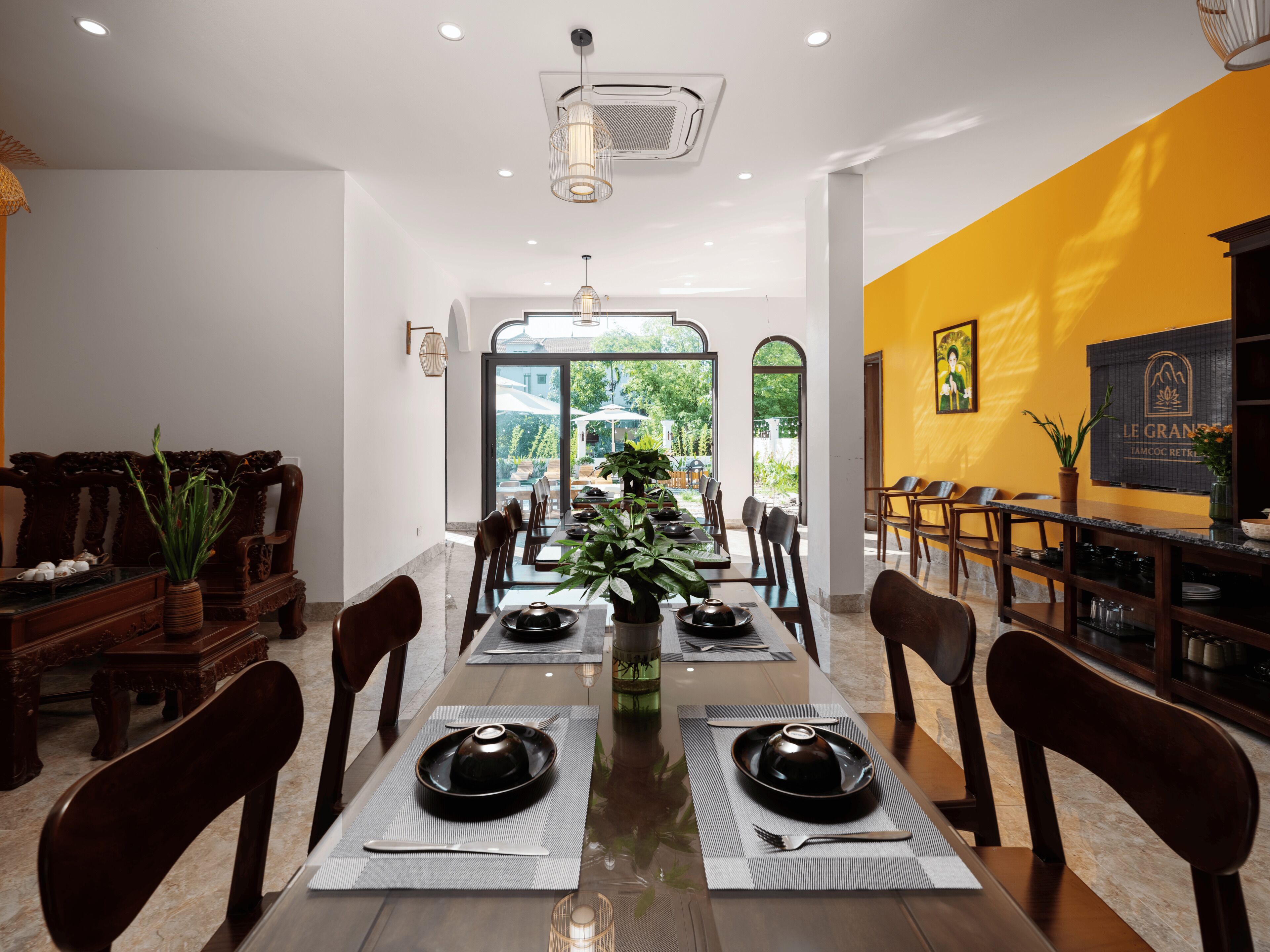A dining area with a long table set for a meal, flanked by wooden chairs and a potted plant.