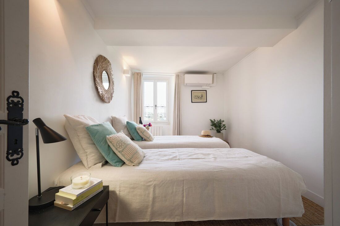 A bedroom with two beds, a bedside table with a lamp, a wall-mounted mirror, and a window with curtains.