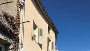 Un edificio de dos pisos con fachada de piedra, un camino de entrada pavimentado y una pequeña área ajardinada con un arbusto y un muro de piedra.