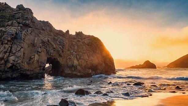 Un paysage côtier avec une formation rocheuse imposante, comprenant une arche naturelle, des vagues déferlant sur une plage de sable, et des collines lointaines sous un ciel au coucher du soleil.