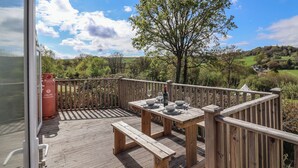 A wooden deck with a table set for dining, overlooking a scenic landscape with hills and a tree. - Willow's Leap (Lyme Regis)
