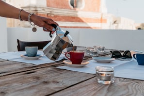 Een hand giet koffie in een rode beker op een houten tafel, met verschillende bekers, schoteltjes, glazen en een kaars.