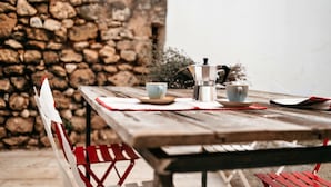A rustic wooden table with a coffee maker, two cups, and a plate, set against a stone wall and red chairs. - Palazzo Pati - Suite Deluxe (Tiggiano)