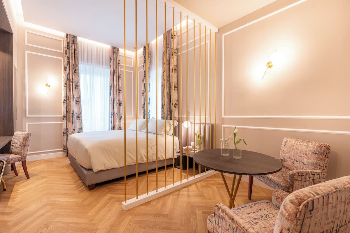 Une chambre d’hôtel comprenant un lit, une table ronde, deux fauteuils à motifs, une petite table d’appoint et un luminaire fixé au mur.