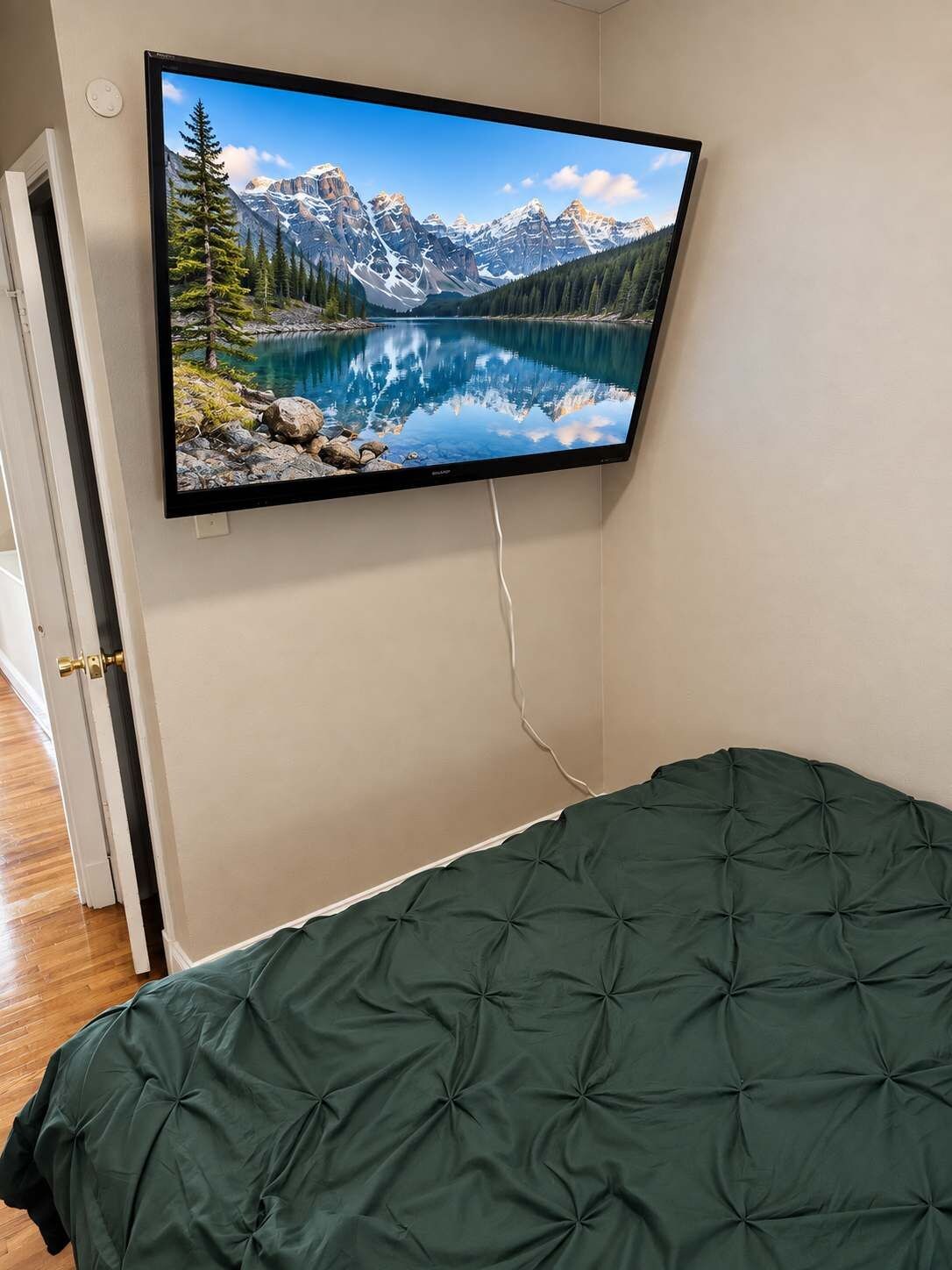 A bedroom with a bed, a wall-mounted TV displaying a mountain landscape, and a door leading to another room.