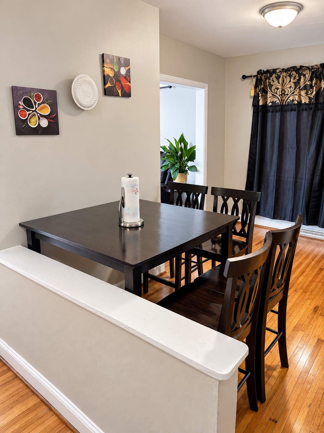 A dining area with a dark wooden table, white bench, and black chairs. There are wall-mounted artworks and a potted plant visible.
