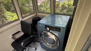 A compact room with a Samsung washing machine, a black office chair, and a view of greenery through large windows. - 3BR King Beds | 85” TV | 4min to A&T & Downtown (Greensboro)