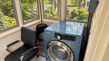 A compact laundry room with a Samsung washing machine, a black office chair, and large windows offering a view of trees and a clear sky.
