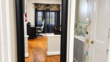 A hallway with a mirror reflecting a room with a desk, chair, and a window with curtains.
