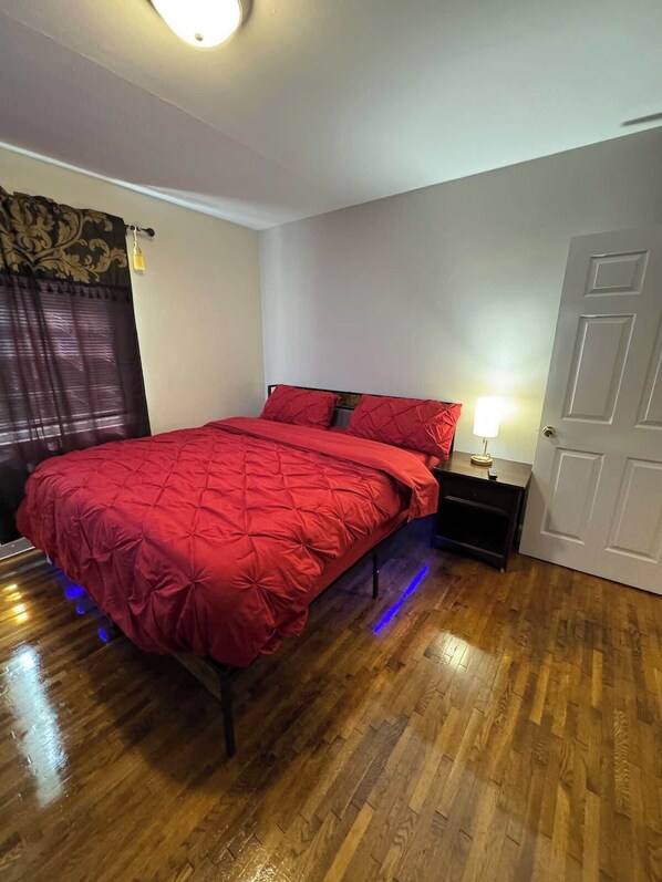 Une chambre à coucher avec une couvre-lit rouge, une table de chevet avec une lampe, une porte et une fenêtre avec des rideaux.