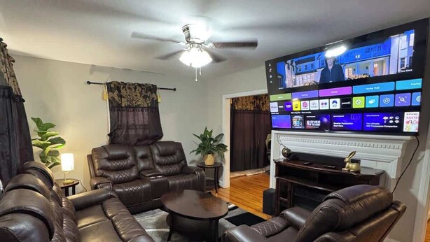 A living room with a leather sofa set, a coffee table, and a flat-screen TV mounted on the wall displaying a home entertainment system interface. - 3BR King Beds | 85” TV | 4min to A&T & Downtown (Greensboro)