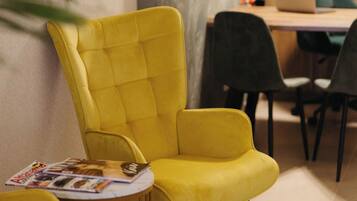 Un sillón amarillo con una revista encima, una mesita de madera y un ordenador portátil sobre un escritorio en una habitación con papel tapiz estampado.