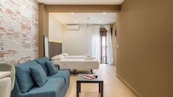 A hotel room with a blue sofa, a small table, a bed with white linens, a brick wall, and a window with curtains.