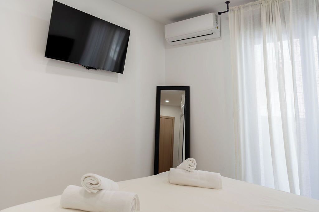 A hotel room with a flat-screen TV mounted on the wall, an air conditioning unit, a large mirror, and two neatly made beds with rolled towels.