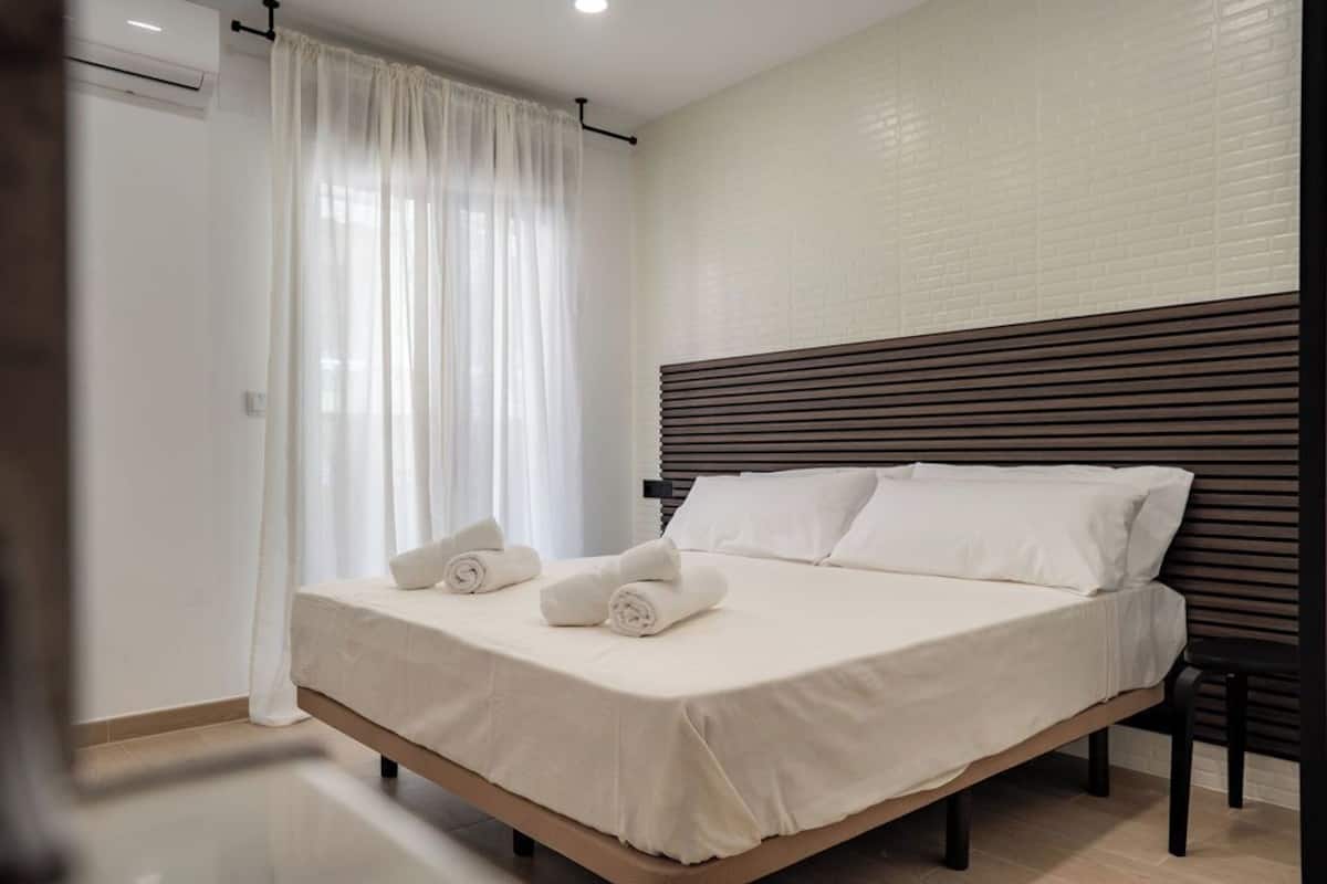 A neatly made bed with white linens and rolled towels, a wooden headboard, and a white brick wall.